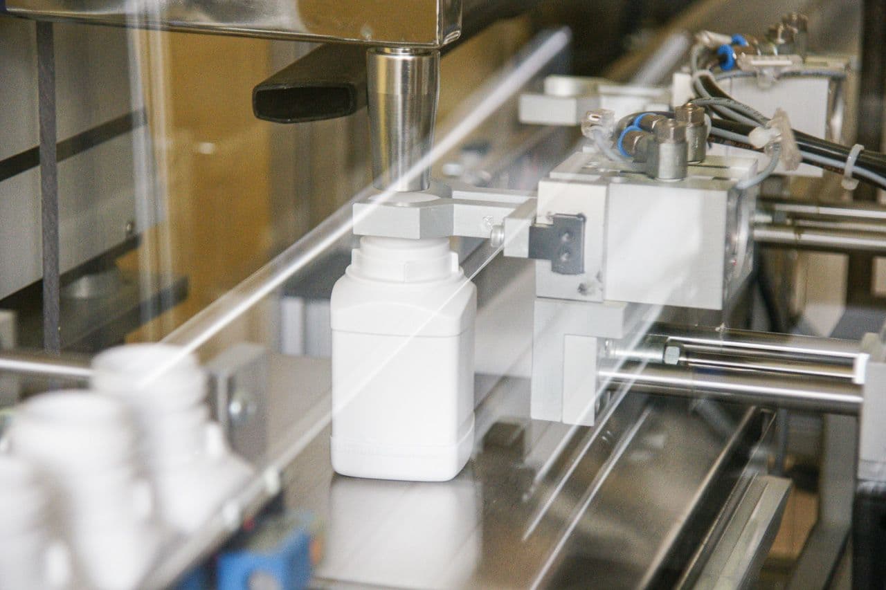 Filling line in operation: a white product bottle under the filling nozzle behind the machine guard.