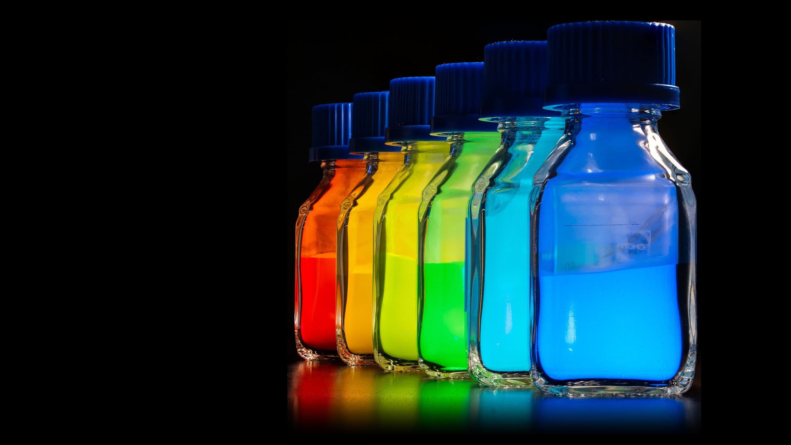 Six laboratory bottles of phosphor powder under excitation, glowing red, orange, yellow, green, cyan and blue against a black background.